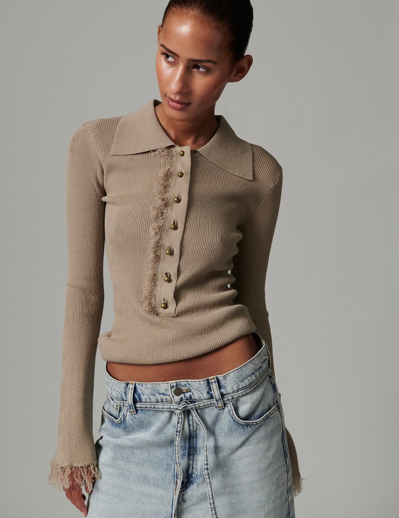 Μπεζ μακρυμάνικο τοπ με ραβδώσεις, αντίθετο γιακά, χρυσά κουμπιά στο μπροστινό μέρος και σκισμένες λεπτομέρειες στο τελείωμα. Συνδυάζεται με ανοιχτόχρωμο σορτς τζιν.
