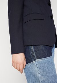 Granatowa marynarka z przeszyciami na ramionach, z klapami z nacięciami i trzema guzikami. Połączona z niebieskimi dżinsami o jasnym odcieniu i kontrastującymi ciemnymi łatami z denimu.