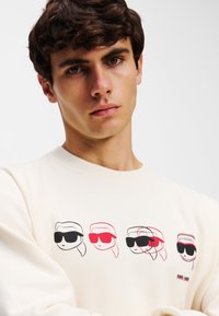 Crème sweatshirt met vier geïllustreerde gezichten met zonnebrillen in zwart en rood. 'KARL LAGERFELD' logo in het rood op de onderste rechterzijde.