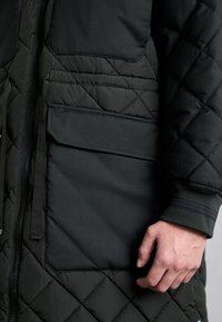 Sötétzöld steppelt dzseki, cipzáras záródással, egy nagy foltokkal ellátott zsebbel és bordázott mandzsettákkal, melyeken két csík található a csukló közelében.