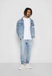 Giacca di jeans con lavaggio azzurro chiaro, due taschini sul petto, t-shirt bianca con logo, jeans azzurri chiari e sneakers bianche. Outfit casual.
