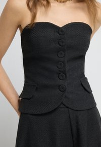 Czarny top bez ramiączek z fakturowanej tkaniny, z zakrzywionym dekoltem, pięcioma dużymi guzikami pokrytymi tkaniną oraz dwoma bocznymi kieszeniami.