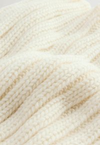 Cremfarbener gestrickter Stoff mit einer strukturierten, groben Musterung und Rippendetails sowie weichen, plüschigen Fasern.