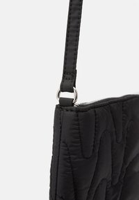 Černá prošívaná taška s elegantním designem, hladkým povrchem, nastavitelým popruhem a stříbrnými kovovými prvky. Zipové uzavření nahoře.