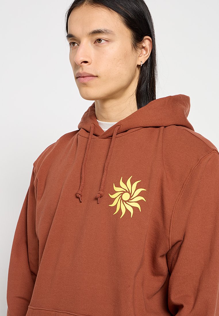 Sweatshirt à capuche marron en coton, avec un graphique en soleil jaune sur la poitrine gauche. Il a une coupe décontractée et confortable.