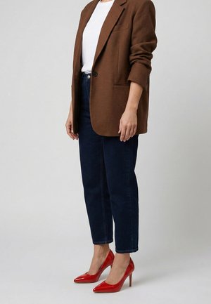 Person iført en brun blazer, hvid skjorte, mørkeblå jeans og røde højhælede sko, der står foran en ensfarvet baggrund.