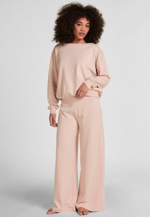 Donna con capelli ricci che indossa una felpa rosa pallido larga e pantaloni a gamba larga, in piedi a piedi nudi contro uno sfondo semplice.