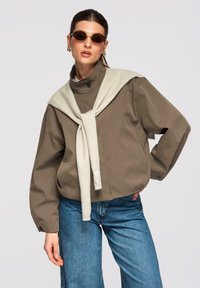 Femme portant des lunettes de soleil rondes, une veste taupe surdimensionnée, un jean bleu et un pull beige clair drapé sur les épaules sur un fond uni.