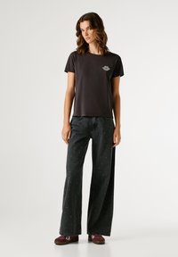 Γυναίκα που στέκεται φορώντας μαύρη μπλούζα με εκτύπωση χείλη, φαρδιά μαύρα τζιν με μικρές κουκίδες και μπορντό παπούτσια σε λευκό φόντο.