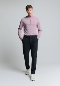 Lichtroze zip-neksweater gecombineerd met zwarte trousers. De sweater heeft een slim fit en een geborduurd logo. Het model draagt witte sneakers.