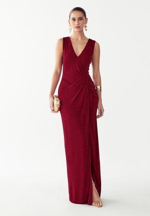 Vrouw draagt een mouwloze dieprode jurk met een zijsplit, houdt een gouden clutch vast en heeft gouden oorbellen, een brede armband en sandalen als accessoires.