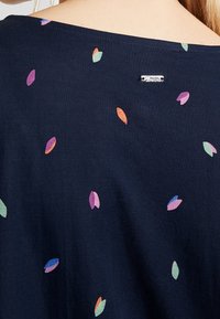 Námorně modrý top s hladkou texturou, se vícero barevnými vzory listů. Obsahuje malý stříbrný detail s logem na zadním výstřihu.