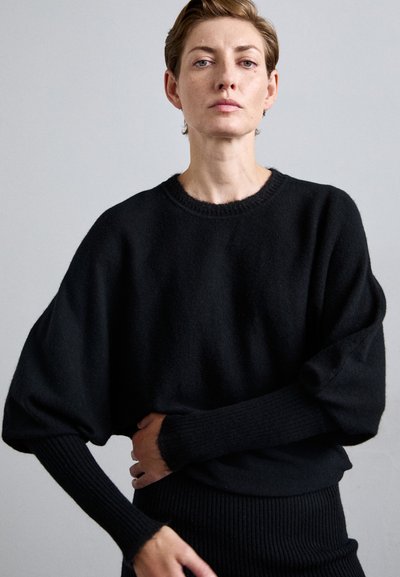 Donna dai capelli corti che indossa un maglione nero largo con maniche lunghe aderenti, in piedi contro uno sfondo grigio chiaro uniforme.