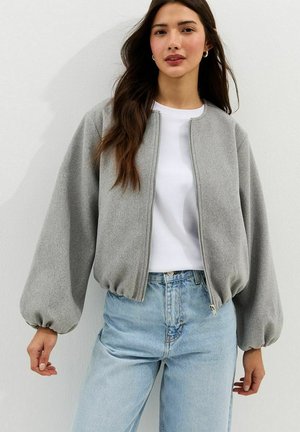 Kobieta w szarej kurtce na zamek z pufiastymi rękawami, na białej koszuli i jasnoniebieskich, wysokich spodniach jeansowych na jednolitym tle.