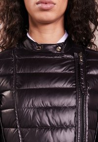Chaqueta de plumas negra con diseño acolchado horizontal, tejido brillante, cuello alto, dos cremalleras y botones de presión en tono dorado.