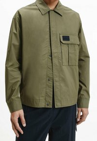 Olivgrüne Jacke aus einem glatten, leichten Stoff. Verfügt über eine Knopfverschluss vorne, einen klassischen Kragen und eine Brusttasche mit Klappe.