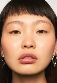 Gros plan d'un visage avec une peau lisse et claire. Les traits incluent des cheveux noirs raides, des yeux marron clair et des boucles d'oreilles géométriques en argent. Les lèvres sont pleines et brillantes.