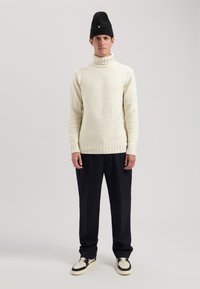 Cremfarbener, grob gestrickter Pullover mit hohem Kragen, schwarze enganliegende Mütze, schwarze Hose und schwarze sowie cremefarbene Sneaker. Einfaches Design und Textur.
