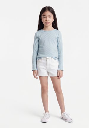 Young girl with long dark hair wearing a light blue long-sleeve top, white shorts, and white sneakers, standing against a plain background.