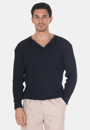 Uomo con capelli scuri che indossa un maglione blu navy e pantaloni beige in piedi con una mano in tasca su sfondo bianco.