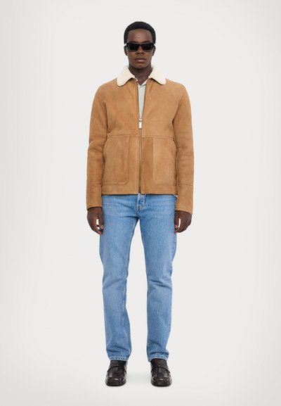 Sundræk jakke i ruskind med shearling-krave, forreste lommer, lynlåslukning, båret over en lys grøn skjorte og blå jeans. Modellen har solbriller på.