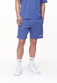 Shorts en coton bleu à coupe décontractée, avec un détail logo. Portés avec des chaussettes de sport blanches et des chaussures blanches pour un look décontracté.