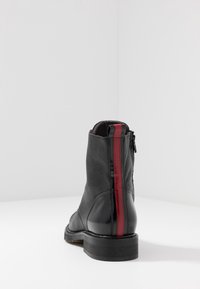 Svart läderankelboot med texturerad yta, röd vertikal rand bak, och sidodragkedja på en vit yta.