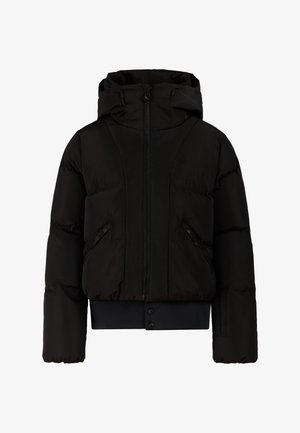 Kind draagt een zwarte winterjas, zwarte handschoenen, een zwarte muts, oranje zonnebril, broek met luipaardprint en zwarte schoenen, staand tegen een lichte achtergrond.