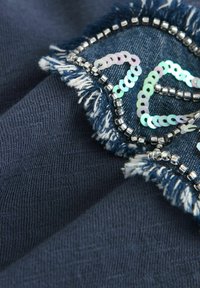 Marinblått tyg med en fransad denimlapp, prydd med silverpärlor och iriserande paljetter, som skapar intrikata mönster och texturer.