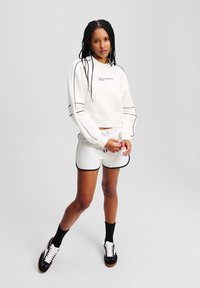 Felpa bianca corta con cuciture nere e dettagli del logo, abbinata a pantaloni corti bianchi con bordi neri. Il modello indossa sneakers nere e calze.