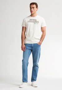 Camiseta blanca de algodón con el texto "PETROL INDUSTRIES & CO" impreso en texto oscuro y verde, combinada con jeans azul claro y zapatillas blancas.