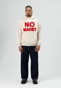 Crèmekleurige hoodie met rode, zachte geborduurde tekst "NO MADE?" en een kangoeroezak, gecombineerd met donkerblauwe wijde jeans en zwarte schoenen.