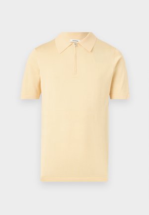 Beige kortærmet polo med krave og en kort lynlås foran, vist på en ensfarvet lys grå baggrund.