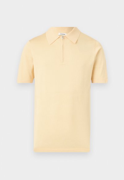 Μπεζ κοντομάνικο πόλο με γιακά και κοντό φερμουάρ μπροστά, απεικονιζόμενο σε απλό ανοιχτό γκρι φόντο.