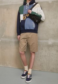 Sweat à capuche patchwork coloré en bleu marine, vert et beige ; short beige ; sac à épaule noir ; chaussettes montant à la cheville ; baskets bleues et blanches.