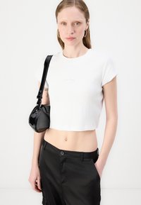 White cropped t-shirt with short sleeves and a subtle logo, paired with a small, shiny black handbag. Black pants with side pockets.