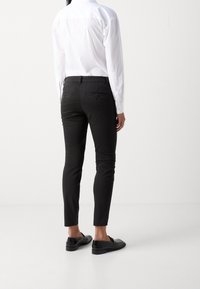 Pantalon noir slim avec une texture lisse, poches arrière et une coupe ajustée, assorti à des mocassins noirs et une chemise blanche à col.