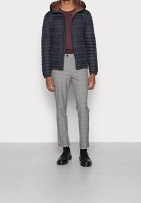 Giacca a vento navy con cappuccio, fodera interna marrone, abbinata a pantaloni a quadri grigi e scarpe nere. Texture liscia e design affusolato.