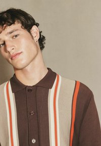 Strick-Polohemd in Braun mit vertikalen Streifen in Beige, Orange und Rot. Mit klassischem Kragen und einer Knopfleiste mit drei Knöpfen.