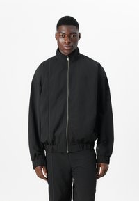 Schwarze Bomberjacke mit Reißverschluss, Stehkragen, elastischen Bündchen und Saum. Sie verfügt über einen strukturierten Stoff und ein minimales Logodesign auf der Brust.