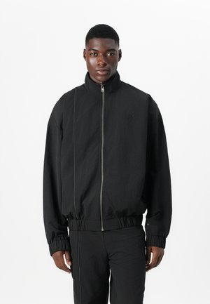 Melna zip-up bomber jakete ar stāvu apkakli, elastīgām aprocēm un jostasvietu. Iezīmējas ar teksturētu audumu un minimālu logotipu uz krūtīm.
