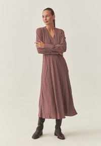 Piros maxi ruha ovális mintával, hosszú ujjú, V-nyakú és középmellmagasságban végződik, barna térdig érő bőr csizmával párosítva.