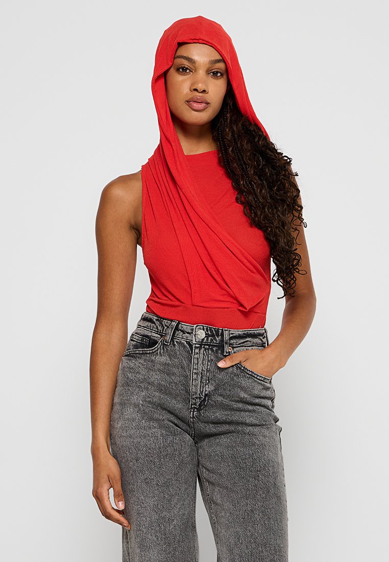 Czerwona bezrękawnik z drapowanym kapturem, wykonany z teksturowanej tkaniny. W zestawieniu z szarymi dżinsami z wysokim stanem, z klasycznym zapięciem na guzik.