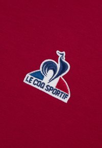 Il tessuto di cotone rosso presenta un logo ricamato di un gallo con accenti blu e bianchi, etichettato "LE COQ SPORTIF" sottostante.