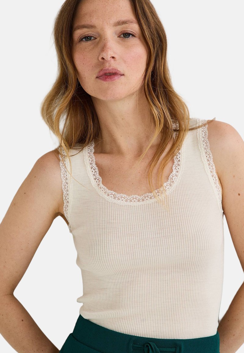 Débardeur beige à côtes avec un volant en dentelle le long de l'encolure et des épaules, associé à une taille haute de couleur vert foncé.