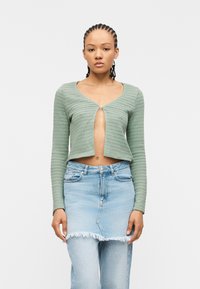 Γυναίκα με κοτσίδες cornrow που φοράει ένα κοντό πράσινο ζακέτα που κλείνει με ένα κουμπί και μια ανοιχτό μπλε τζι ν φούστα πάνω από τζιν παντελόνι.