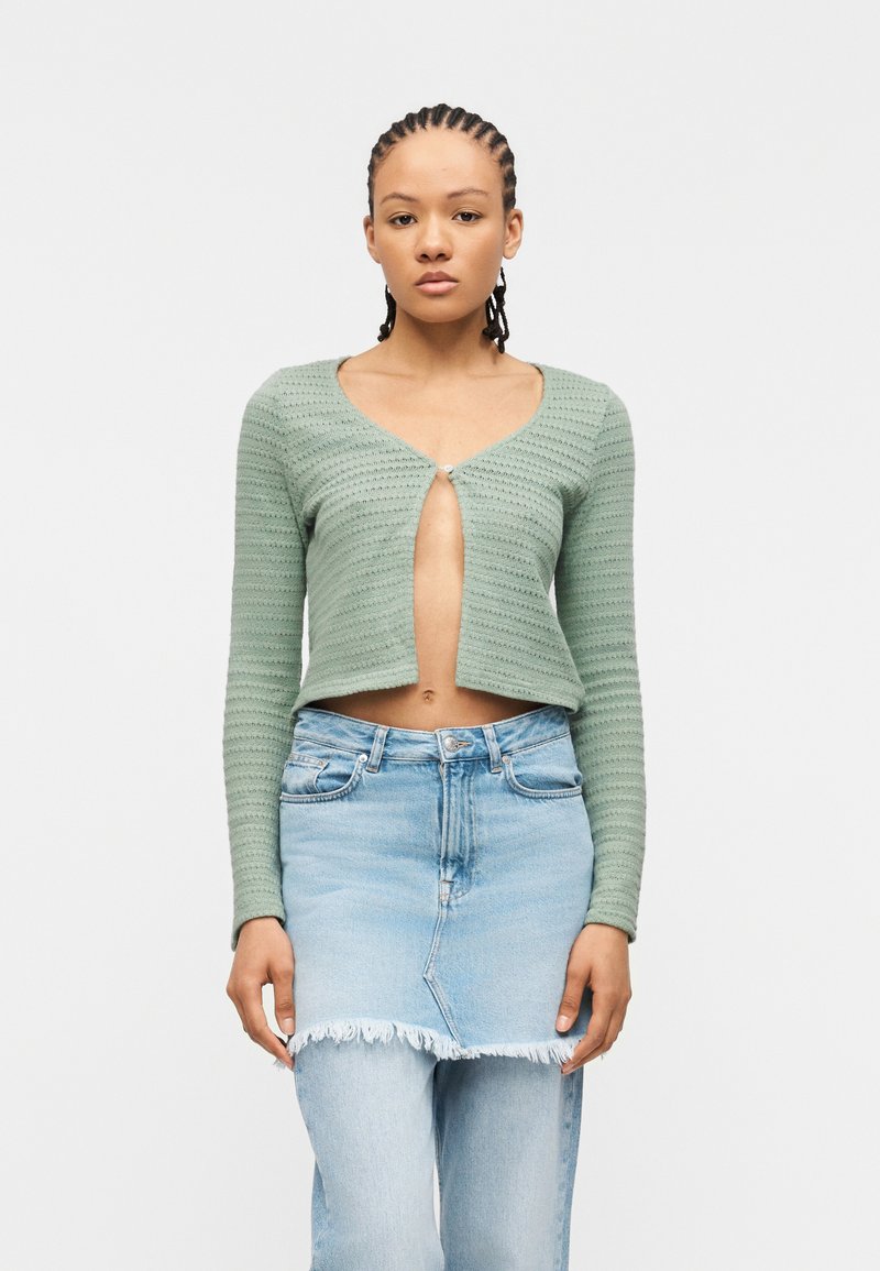 Γυναίκα με κοτσίδες cornrow που φοράει ένα κοντό πράσινο ζακέτα που κλείνει με ένα κουμπί και μια ανοιχτό μπλε τζιν φούστα πάνω από τζιν παντελόνι.