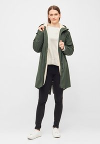 Groene waterdichte parka met een capuchon gevoerd met nepbont, sluitingen aan de voorkant en een taille met trekkoord, gedragen over een lichte trui en zwarte jeans.