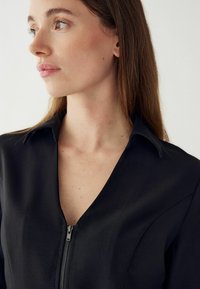 Melna kleita ar apkakli un V-veida izgriezumu, priekšā ar sudraba rāvējslēdzēja detaļu. Gluda auduma un pielāgota forma, kas akcentē izgriezumu.