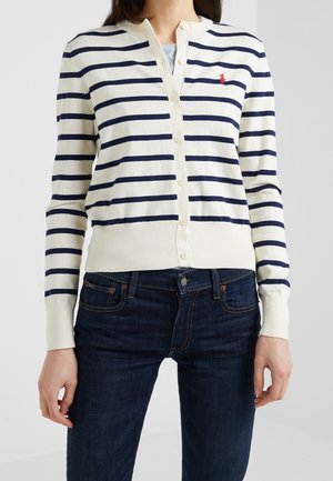 Femme portant un gilet blanc à rayures horizontales bleu marine avec un logo rouge, associée à un jean bleu foncé, debout devant un fond uni.